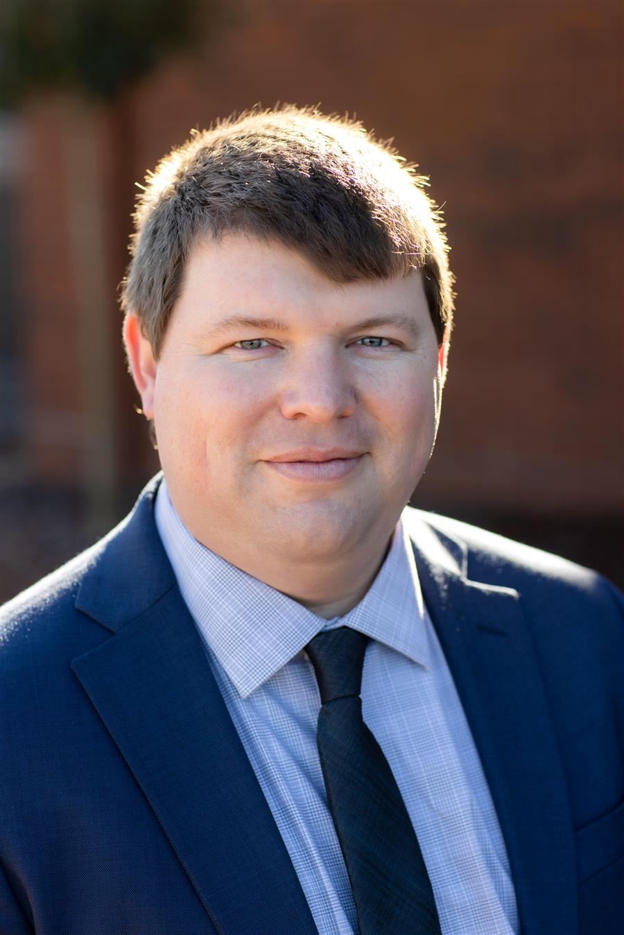  Kyle Gregg in a blue suit and tie with a blurred brick wall background.